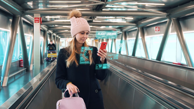 Eine junge Frau mit Gepäck fährt ein einem Flughafengebäude auf einem Fußgängerfließband und bedient ihr Smartphone
