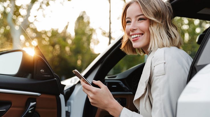 Laughing woman sits on driver's seat of open car and operates her smartphone