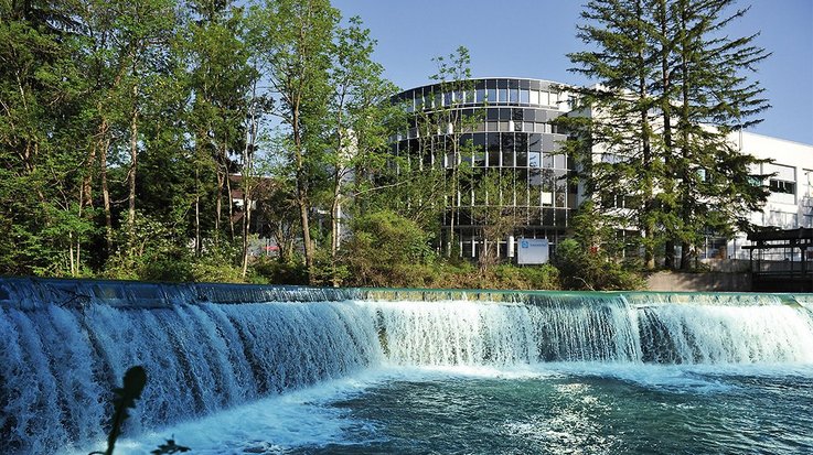 Ein kleiner Wasserfall an einem breiten Gewässer, im Hintergrund ein Bürogebäude