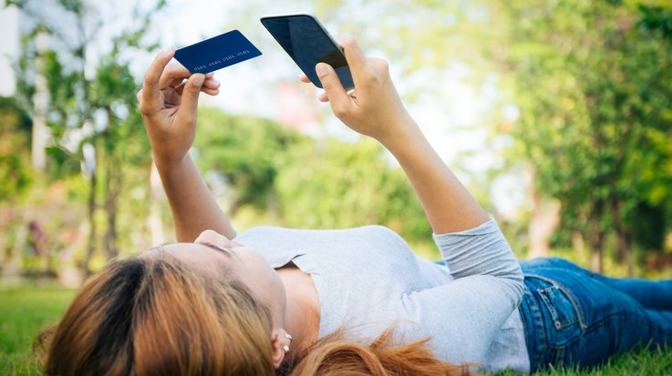 A woman lies on a meadow holding a credit card in one hand and a smartphone in the other