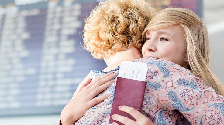 Two women embrace on an airport terminal, one holds her passport in her hand