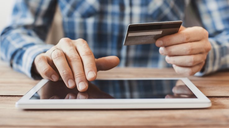 Ein Mann sitzt an einem Tisch und bedient ein Tablet, während er in einer Hand eine Kreditkarte hält