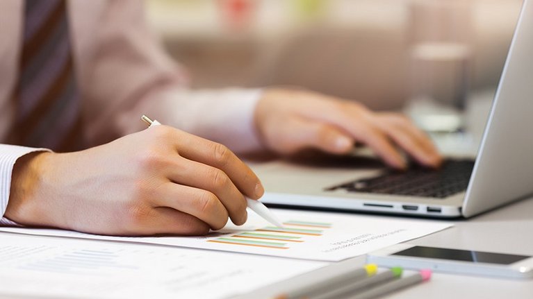 Man sitting at desk, holding pen in hand, has a printed graphic in front of him and is typing on his laptop