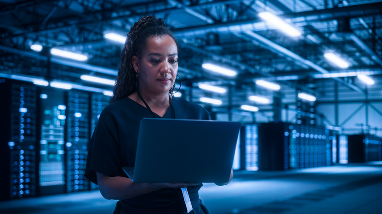 Woman standing in front of a server farm while holding and looking at a laptop