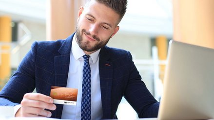 A smiling businessman sits in front of his laptop and holds a credit card in his hand