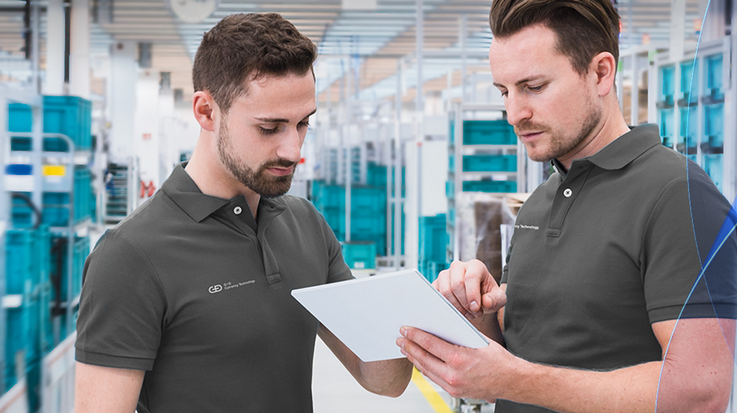 Two G+D employees in polo shirts discuss a clipboard, NotaTracc trays are in the background