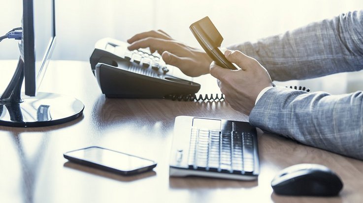 Man sits at a desk, has a telephone receiver in his hand and types in a number