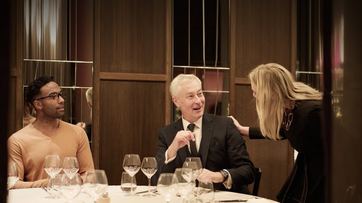 Drei gut gekleidete Personen treffen sich an einem feinen gedeckten Tisch in einem gehobenen Restaurant