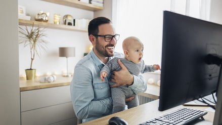 A man sits laughing with a baby in his arms in front of a computer screen