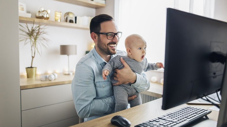 A man sits laughing with a baby in his arms in front of a computer screen