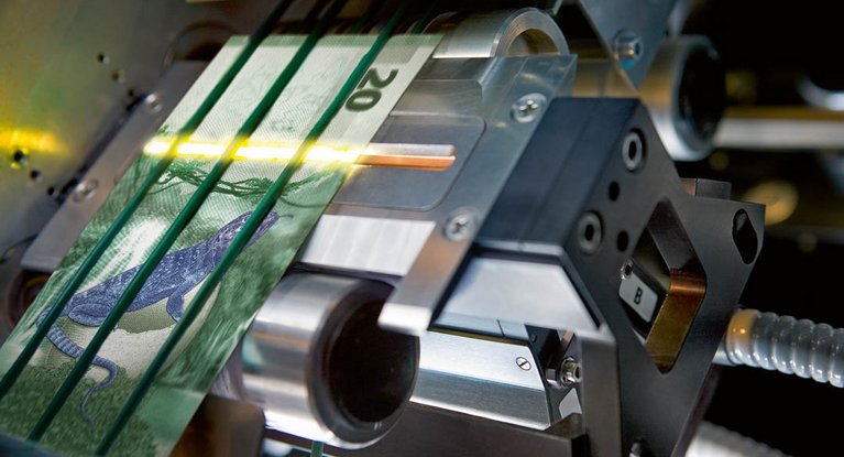 Close-up image of a banknote processing machine with ultrasonic sensors and a sample note