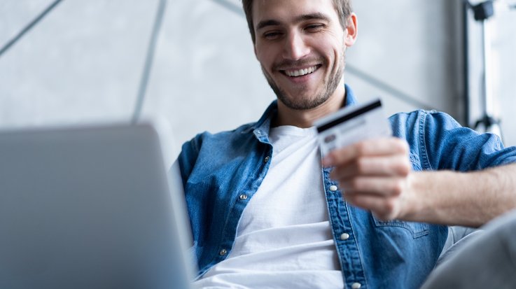 A smiling young man has an open laptop on his lap and holds a credit card in his hand