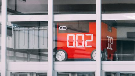 Behind the window pane of a high-rise building stands a red machine on wheels with the G+D Currency Technology logo 