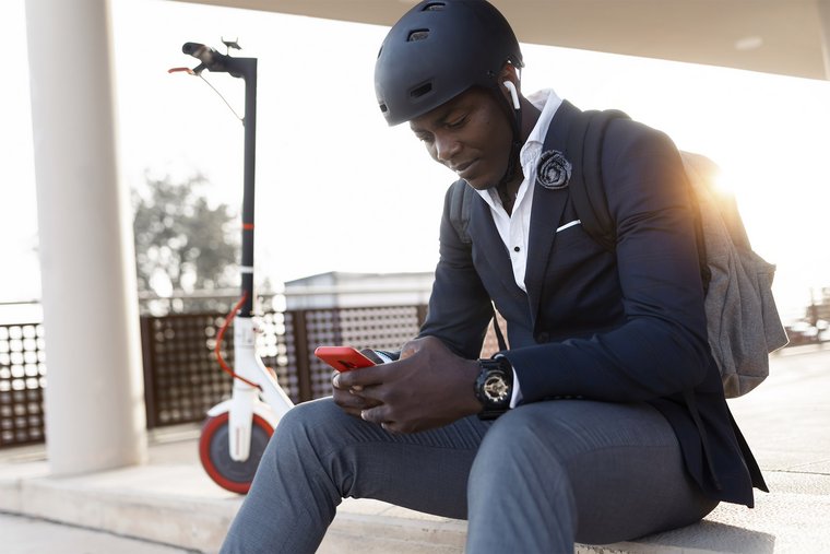 Junger Geschäftsmann mit Roller sitzt auf Treppe bei Sonnenuntergang mit Handy und Kopfhörern