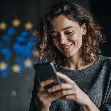 Eine lächelnde junge Frau, die auf ihr Smartphone schaut, vor einem unscharfen, hellen Hintergrund mit EU-Motiven.