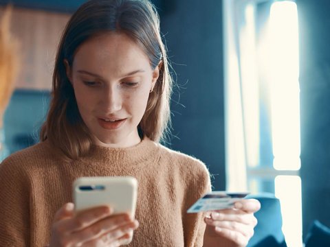 A woman sits in the kitchen holding a smartphone in one hand and a credit card in the other