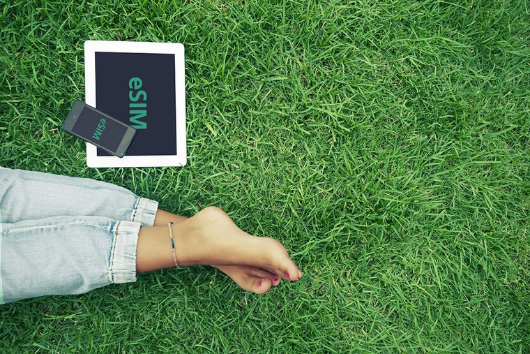 Bare female feet on a lawn, next to them are a tablet and a smartphone showing 'eSIM'.