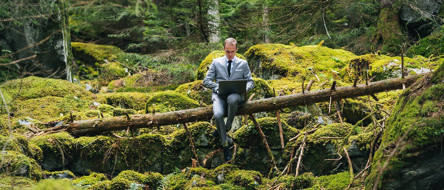Bild eines Mannes der im Wald mit grüner Umgebung an seinen Laptop sitzt und arbeitet.