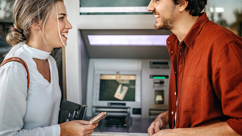 Ein Paar steht lachend vor einem Geldautomaten