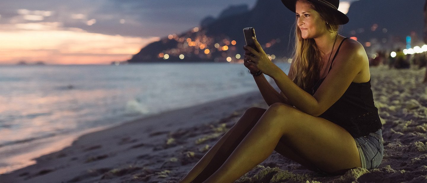 Junge Frau sitzt nachts am Strand und schaut auf ihr Mobiltelefon