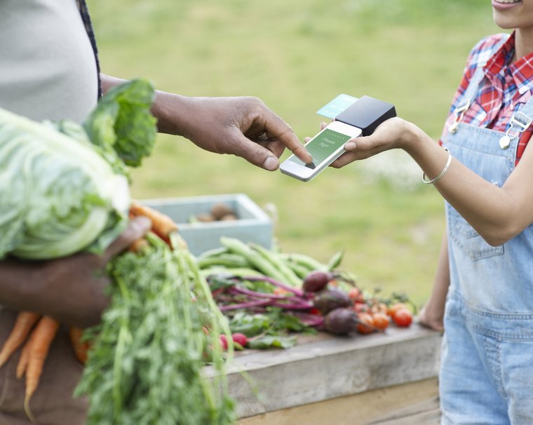 A man pays a woman on a market  for some vegetables using a mobile payment device.