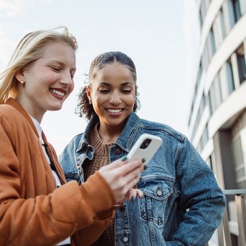 Two young women looking at a white smartphone with futuristic building in the background.