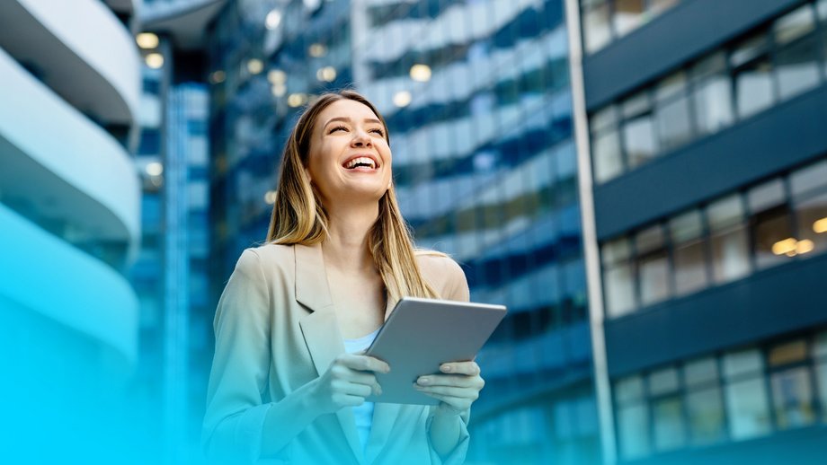 a smiling woman holding a tablet in front of high buildings