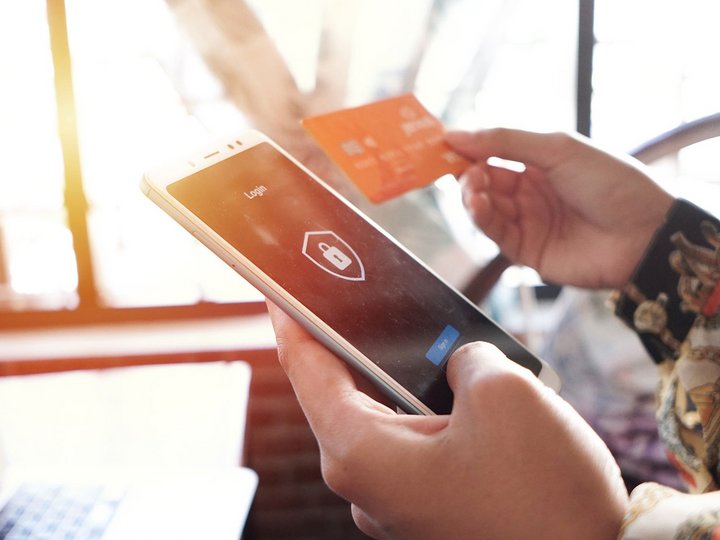 Die linke Hand hält ein Smartphone, das eine Zahlung auf einem Login-Bildschirm verifiziert, die rechte Hand hält eine Kreditkarte.