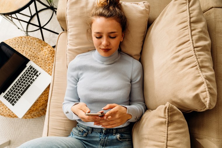 Junge Frau liegt entspannt auf der Couch mit dem Smartphone in der Hand