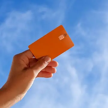 A man's hand holds an organ-colored credit card up against the blue sky.