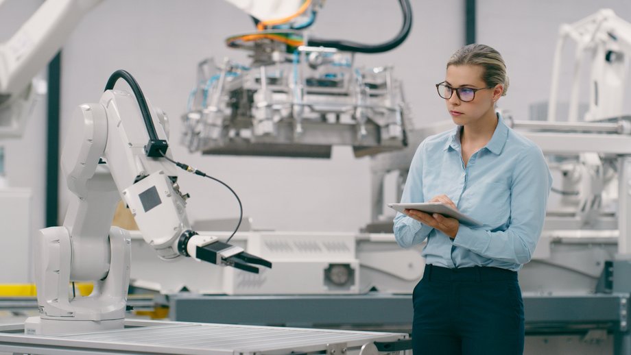 Eine Ingenieurin überwacht einen Roboterarm in einer modernen Fabrikumgebung, während sie ein digitales Tablet hält. 