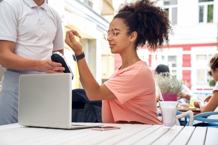 Eine Frau sitzt mit einem Laptop in einem Café und bezahlt kontaktlos mit ihrer Smartwatch